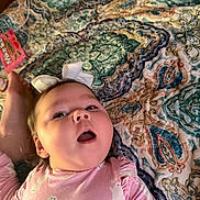 Victoria is registered to the contest to win money with this photo: baby, child, infant, portrait, lying_down, bed, bedspread, bow, pink_clothing, onesie, face, eyes, open_mouth, smiling, indoor, home, textile, patterned_background, close_up, cute