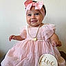 baby, child, girl, pink_dress, headband, bow, pearl_necklace, smiling, portrait, indoor, sign, celebration, milestone, cute, happy, infant, person, clothing, sitting, young
