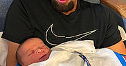 Dalton is registered to the contest to win money with this photo: man, newborn, baby, blanket, holding, sleeping, chair, indoor, portrait, love, family, tender, gentle, person, curly_hair, facial_hair, black_shirt, resting, close_up, caring
