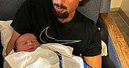 Dalton is registered to the contest to win money with this photo: baby, black_tshirt, blanket, chair, comfort, curly_hair, facial_hair, family, holding, human, indoor, love, man, newborn, parent, portrait, relaxation, resting, sleeping, tenderness