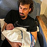 man, newborn, baby, blanket, curly_hair, facial_hair, black_tshirt, chair, indoor, resting, holding, sleeping, portrait, human, parent, family, comfort, love, tenderness, relaxation