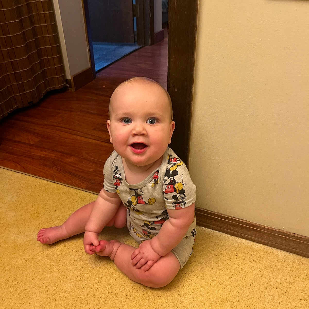 Declan joined the competition — help win amazing prizes! baby, barefoot, child, cute, doorway, floor, happy, home, indoor, infant, mickey_mouse, onesie, person, plush, room, sitting, smiling, wall, window, wooden_floor