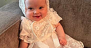 Leia is registered to the contest to win money with this photo: baby, child, smile, lace_dress, bonnet, couch, indoor, happy, sitting, infant, cute, portrait, clothing, fabric, homedecor, person, foot, hand, wall, plant