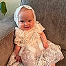 baby, child, smile, lace_dress, bonnet, couch, indoor, happy, sitting, infant, cute, portrait, clothing, fabric, homedecor, person, foot, hand, wall, plant