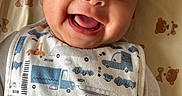 Ibrahim is registered to the contest to win money with this photo: baby, smile, infant, bib, blanket, teddy_bear, clothing, hand, face, happy, cute, child, lying_down, indoors, person, portrait, skin, eyes, mouth, baby_garment