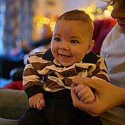 Elijah is registered to the contest to win money with this photo: adult, baby, blurred_background, casual_clothing, child, couch, cozy, cute, face, hand, happy, holding_hand, indoor, infant, person, portrait, sitting, smile, striped_shirt, warm_lighting