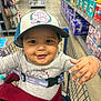 toddler, child, shopping_cart, hat, smile, baby_products, aisle, store, clothing, safety_belt, hand, face, person, retail, sitting, cute, cap, happy, indoor, playful
