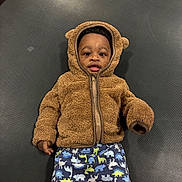 Blessan is registered to the contest to win money with this photo: baby, child, hoodie, brown, fuzzy, dinosaur_pants, lying_down, cute, smiling, face, infant, clothing, texture, black_background, playful, curious, person, indoor, young_child, zipper