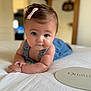 baby, pink_bow, blue_eyes, baby_clothing, bed, white_bedspread, indoor, portrait, child, cute, infant, hands, face, skin, hair, room, blurred_background, curious_expression, lying_down, soft_lighting