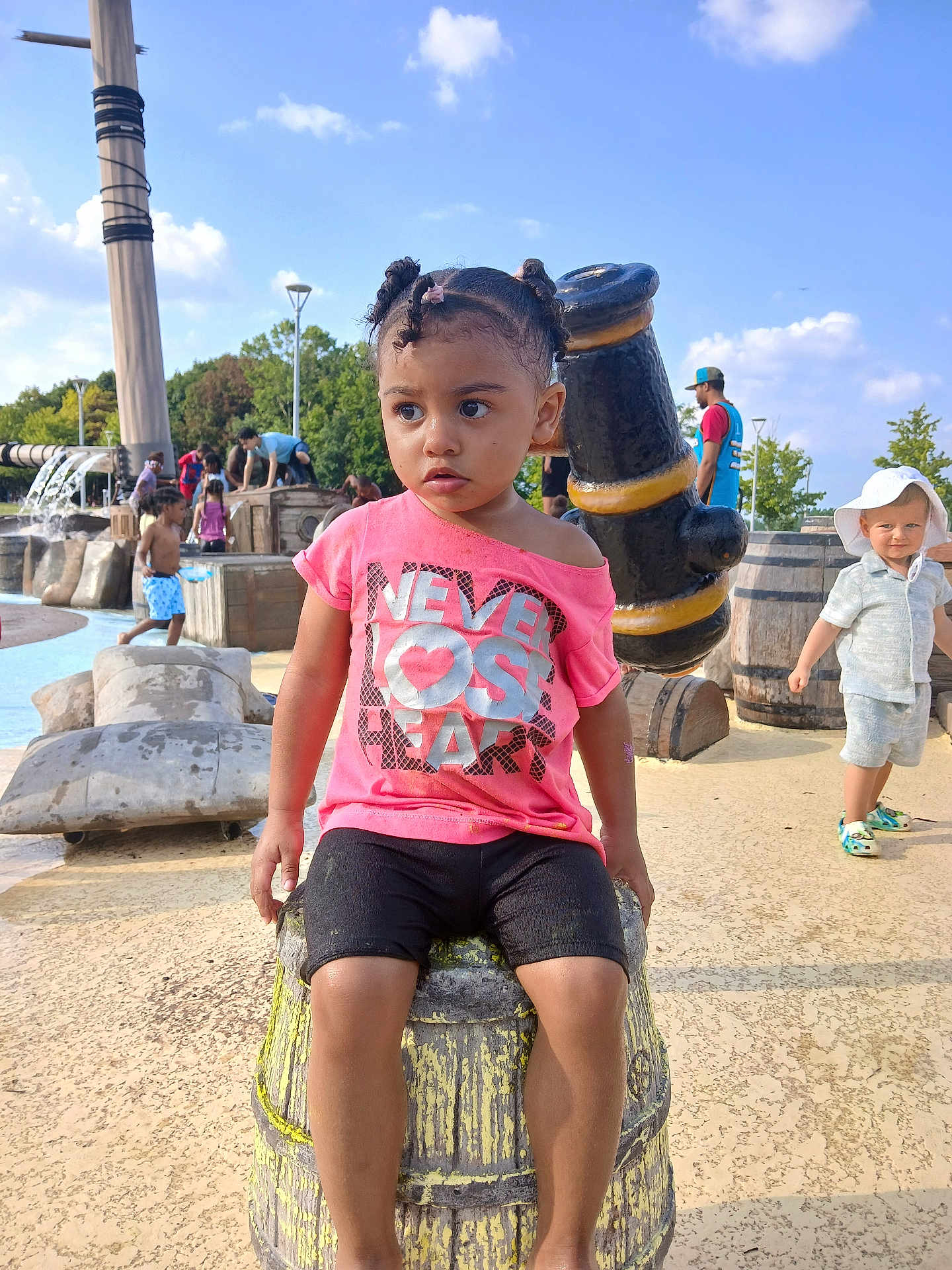 Christiana joined the competition — help win amazing prizes! child, toddler, outdoor, playground, water_park, barrel, pink_shirt, shorts, people, sunny, daytime, trees, blue_sky, play, hat, footwear, expression, park, fun, summer