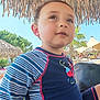child, boy, swimwear, blue, striped, outdoor, sunny, umbrella, thatched_roof, tropical, palm_tree, beach, poolside, curious, young, casual, daylight, vacation, relaxation, portrait