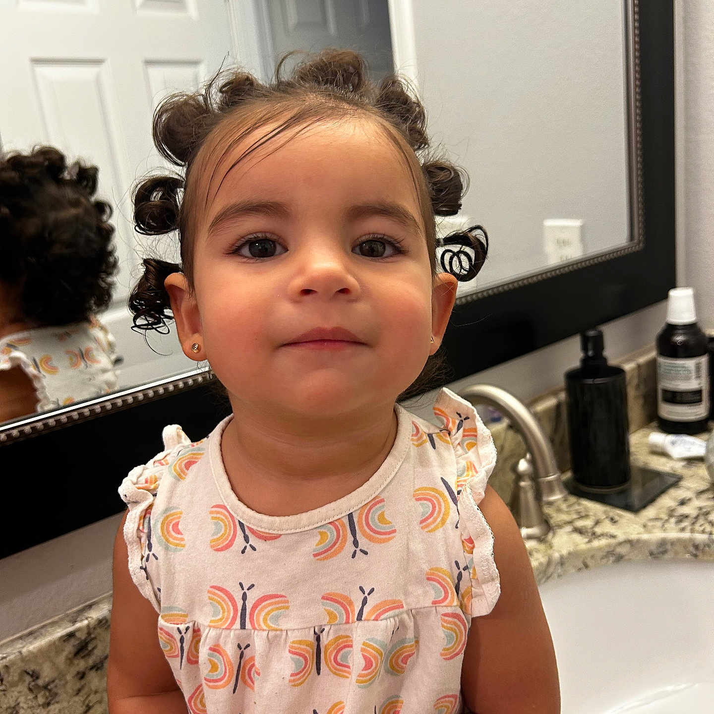 Natalia is registered to the contest to win money with this photo: bathroom, butterfly_pattern, casual, child, countertop, curly_hair, cute, hair_buns, home, indoor, mirror, patterned_clothing, person, portrait, reflection, sink, sitting, smiling, toddler, young_child