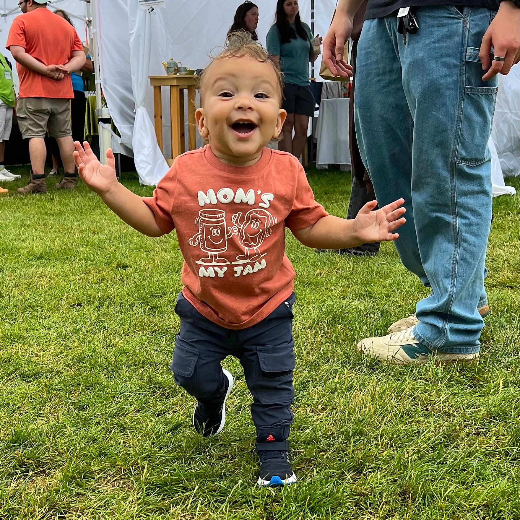 Luca is registered to the contest to win money with this photo: adult, casual_clothing, child, daylight, event, family, festival, fun, grass, greenery, happy, outdoor, people, running, shirt, shorts, smiling, sneakers, tent, toddler