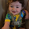 toddler, baby, child, curly_hair, smiling, colorful_clothing, onesie, baby_seat, harness, happy, indoor, wood_floor, person, cute, infant, young_child, portrait, seat_belt, reaching, playful