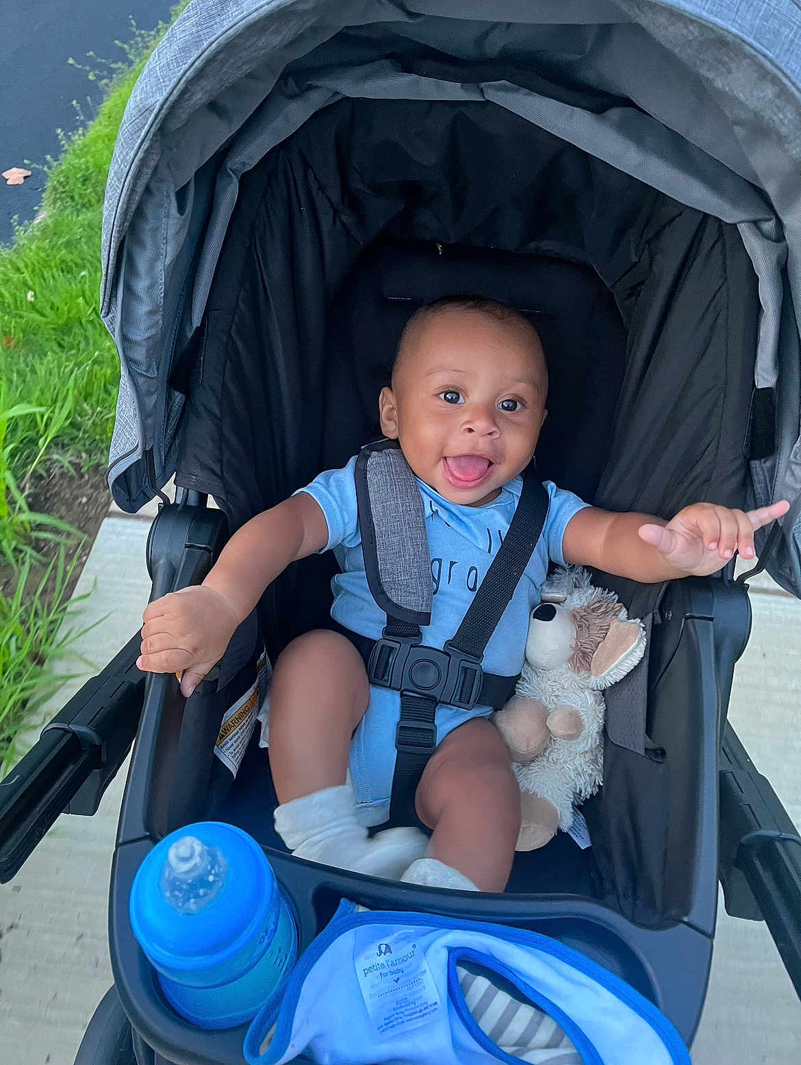 Carlos joined the competition — help win amazing prizes! baby, stroller, blue_onesie, socks, plush_toy, baby_bottle, outdoor, sidewalk, grass, happy, smiling, child, infant, seatbelt, black_stroller, person, cute, portrait, baby_clothing, playful