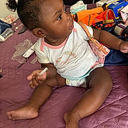 Aylani is registered to the contest to win money with this photo: baby, child, infant, bed, purple_bedspread, clothing, purse, earrings, curly_hair, diaper, indoor, person, sitting, hand, foot, face, cute, casual, clutter, toy