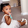 toddler, child, white_dress, carpet, toothbrush, sitting, indoors, curly_hair, shoes, playful, cute, home, floor, window_blinds, furniture, portrait, young_child, baby, person, smiling