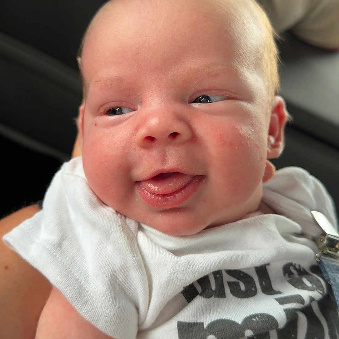 Bo Bradley is registered to the contest to win money with this photo: arm, baby, child, closeup, clothing, cute, face, happy, head, indoor, infant, newborn, person, portrait, seat, sitting, skin, smiling, tongue_out, white_shirt