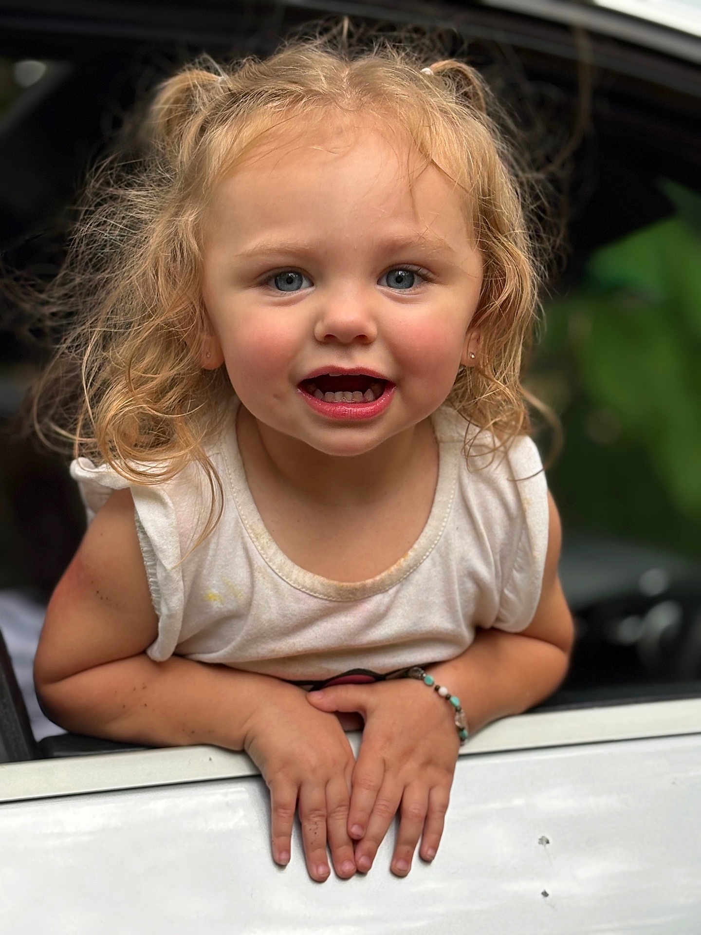 Maelynn joined the competition — help win amazing prizes! toddler, child, blonde_hair, blue_eyes, bracelet, car_window, white_shirt, smiling, hands, portrait, outdoor, curly_hair, cute, young_child, person, casual_clothing, natural_light, happy, closeup, expression