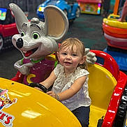 Maelynn joined the competition — help win amazing prizes! child, toddler, smiling, indoor, play_area, kiddie_ride, toy_car, yellow_car, red_car, mouse_mascot, amusement_center, fun, happy, steering_wheel, short_sleeves, bracelet, colorful, entertainment, young_child, casual_clothing