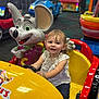 child, toddler, smiling, indoor, play_area, kiddie_ride, toy_car, yellow_car, red_car, mouse_mascot, amusement_center, fun, happy, steering_wheel, short_sleeves, bracelet, colorful, entertainment, young_child, casual_clothing