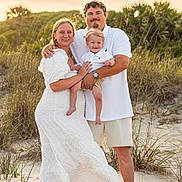 Hardin is registered to the contest to win money with this photo: family, child, toddler, beach, sand, sunset, grass, nature, smile, happy, outdoor, portrait, barefoot, casual_clothing, white_dress, shorts, father, mother, holding, together