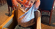 Hardin is registered to the contest to win money with this photo: child, toddler, high_chair, smiling, restaurant, indoor, table, chair, person, casual_clothing, shorts, polo_shirt, blond_hair, happy, carpet, window, lighting, waiter, umbrella, plant