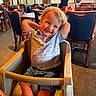 child, toddler, high_chair, smiling, restaurant, indoor, table, chair, person, casual_clothing, shorts, polo_shirt, blond_hair, happy, carpet, window, lighting, waiter, umbrella, plant