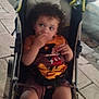 Kaïs participe au concours pour gagner de l'argent avec cette photo : toddler, child, stroller, curly_hair, orange_shirt, mickey_mouse, snack, outdoor, pavement, sidewalk, street, curb, evening, casual_clothing, person, seatbelt, baby, holding_food, looking_away, urban