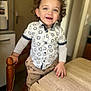 toddler, child, curly_hair, smiling, standing, shirt, pants, indoor, table, chair, wooden, happy, playful, person, footwear, bright_eyes, grip, home, casual_clothing, young