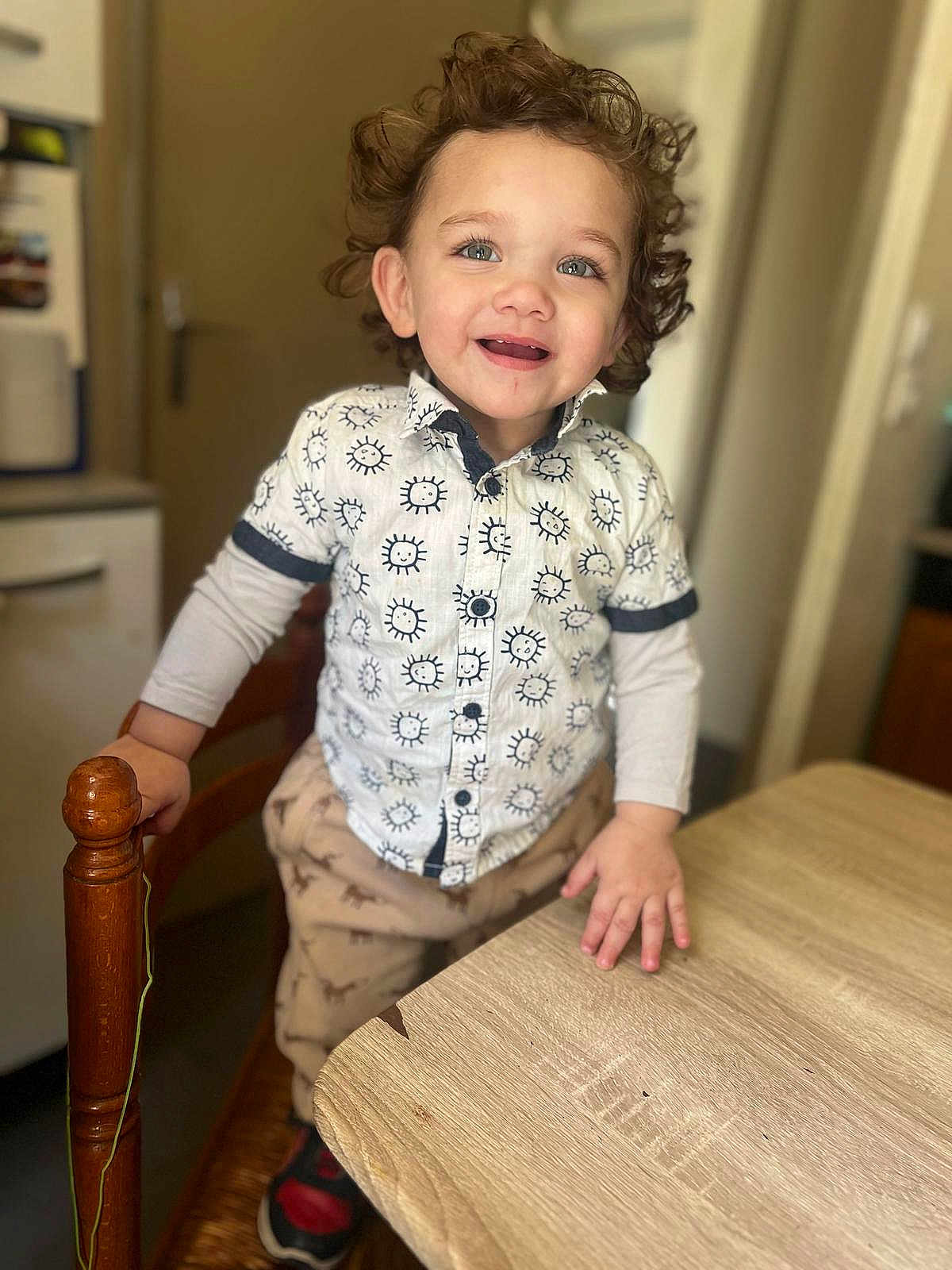 Kaïs participe au concours pour gagner de l'argent avec cette photo : toddler, child, curly_hair, smiling, standing, shirt, pants, indoor, table, chair, wooden, happy, playful, person, footwear, bright_eyes, grip, home, casual_clothing, young