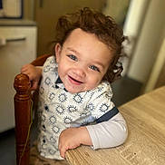 Kaïs a rejoint le concours — aidez-le/la à gagner de superbes lots ! toddler, child, smiling, curly_hair, indoor, table, wooden_table, shirt, patterned_clothing, happy, face, person, sitting, furniture, hand, blue_eyes, cute, young_child, casual_clothing, home