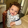 toddler, child, smiling, curly_hair, indoor, table, wooden_table, shirt, patterned_clothing, happy, face, person, sitting, furniture, hand, blue_eyes, cute, young_child, casual_clothing, home