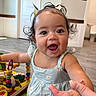 Kahlani is registered to the contest to win money with this photo: baby, child, girl, curly_hair, pigtails, blue_dress, toy, wooden_toy, playroom, happy, smiling, indoor, floor, hand, person, face, eyes, fingers, cute, infant