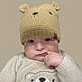 baby, child, knitted_hat, bear_hat, beige_clothing, sweater, cute, infant, face, hand, finger_in_mouth, portrait, expression, soft_lighting, neutral_background, warm_clothing, child_fashion, adorable, close_up, young_child