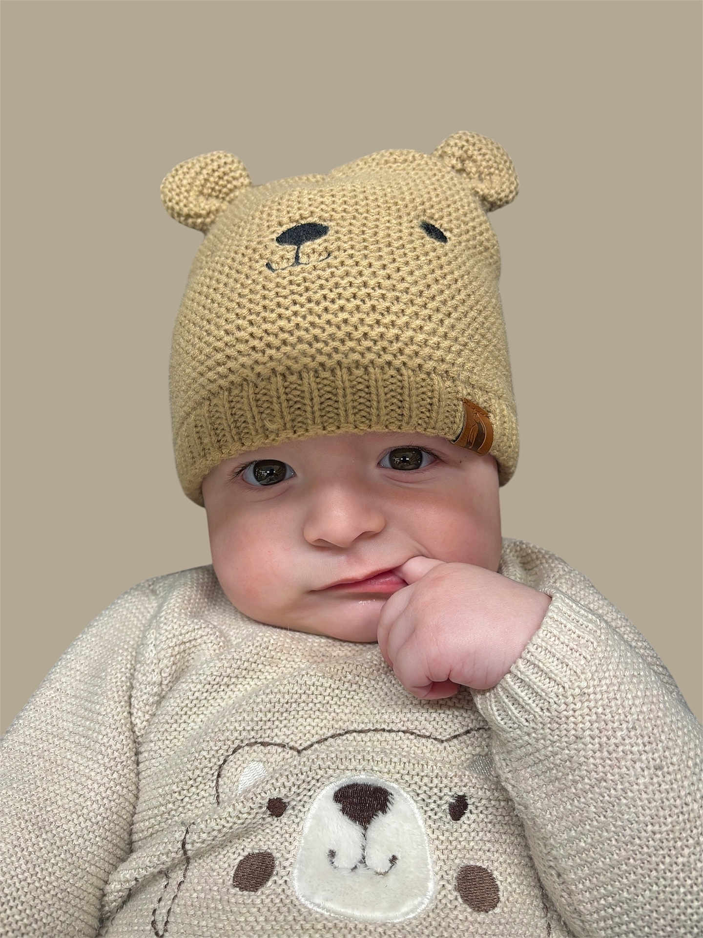 Rome Alexander is registered to the contest to win money with this photo: baby, child, knitted_hat, bear_hat, beige_clothing, sweater, cute, infant, face, hand, finger_in_mouth, portrait, expression, soft_lighting, neutral_background, warm_clothing, child_fashion, adorable, close_up, young_child