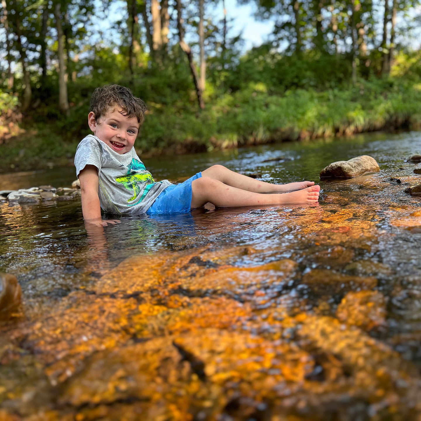 Lane joined the competition — help win amazing prizes! boy, child, curly_hair, forest, happy, nature, outdoor, playful, reflection, rocks, shallow_water, shorts, sitting, smiling, stream, summer, sunlight, trees, tshirt, water