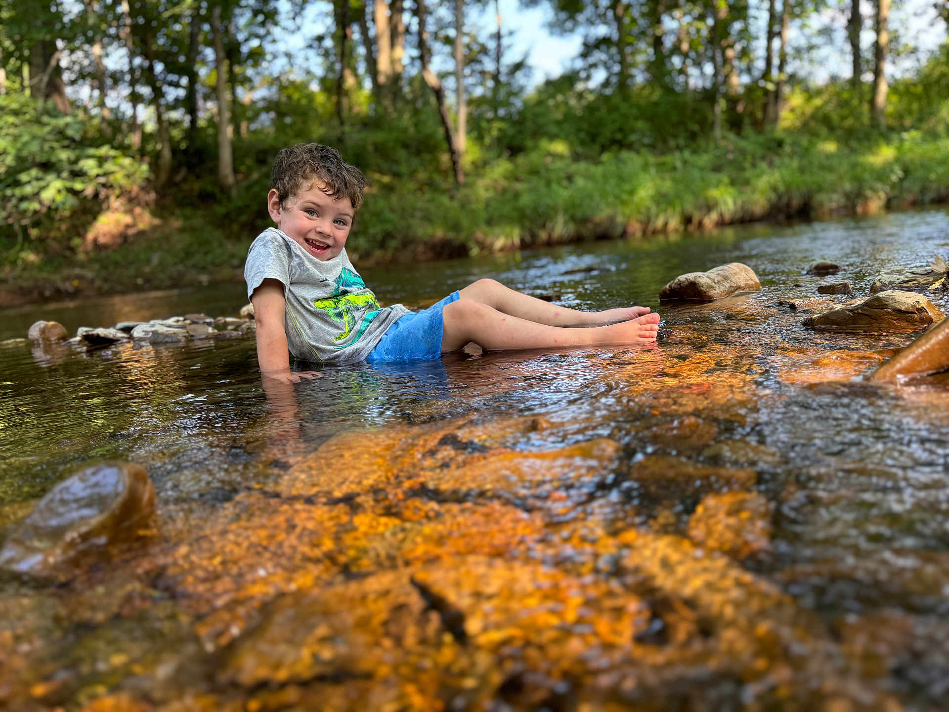 Lane joined the competition — help win amazing prizes! child, boy, water, stream, rocks, forest, nature, outdoor, summer, happy, smiling, shorts, tshirt, curly_hair, sunlight, reflection, playful, sitting, shallow_water, trees