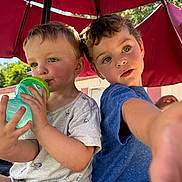 Lane is registered to the contest to win money with this photo: child, children, boy, umbrella, outdoor, daylight, greenery, drink, bottle, hand, curious, playful, red_umbrella, short_sleeves, casual_clothing, portrait, sunlight, nature, sitting, face