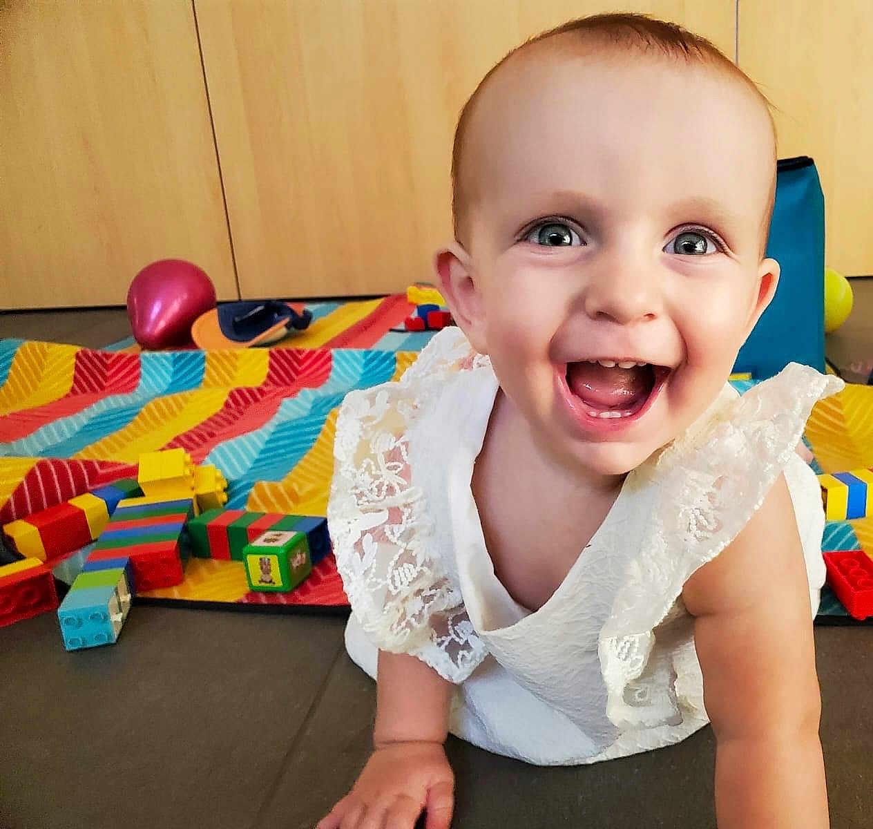 Lison participe au concours pour gagner de l'argent avec cette photo : baby, child, smiling, crawling, white_dress, lace, indoor, play_mat, colorful_blocks, toys, happy, blue_eyes, face, person, cute, floor, wooden_cabinet, playtime, childhood, bright