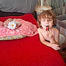 child, toddler, indoors, bedroom, bed, red_blanket, pillow, flower, snack, eating, messy_hair, portrait, sitting, lying_down, crib, fan, furniture, wall, home, candid