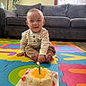 Liam is registered to the contest to win money with this photo: baby, birthday_cake, candle, celebration, child, clothing, colorful, couch, cute, foam_mat, foot, happy, indoor, living_room, pants, plant, portrait, sitting, smiling, striped_shirt