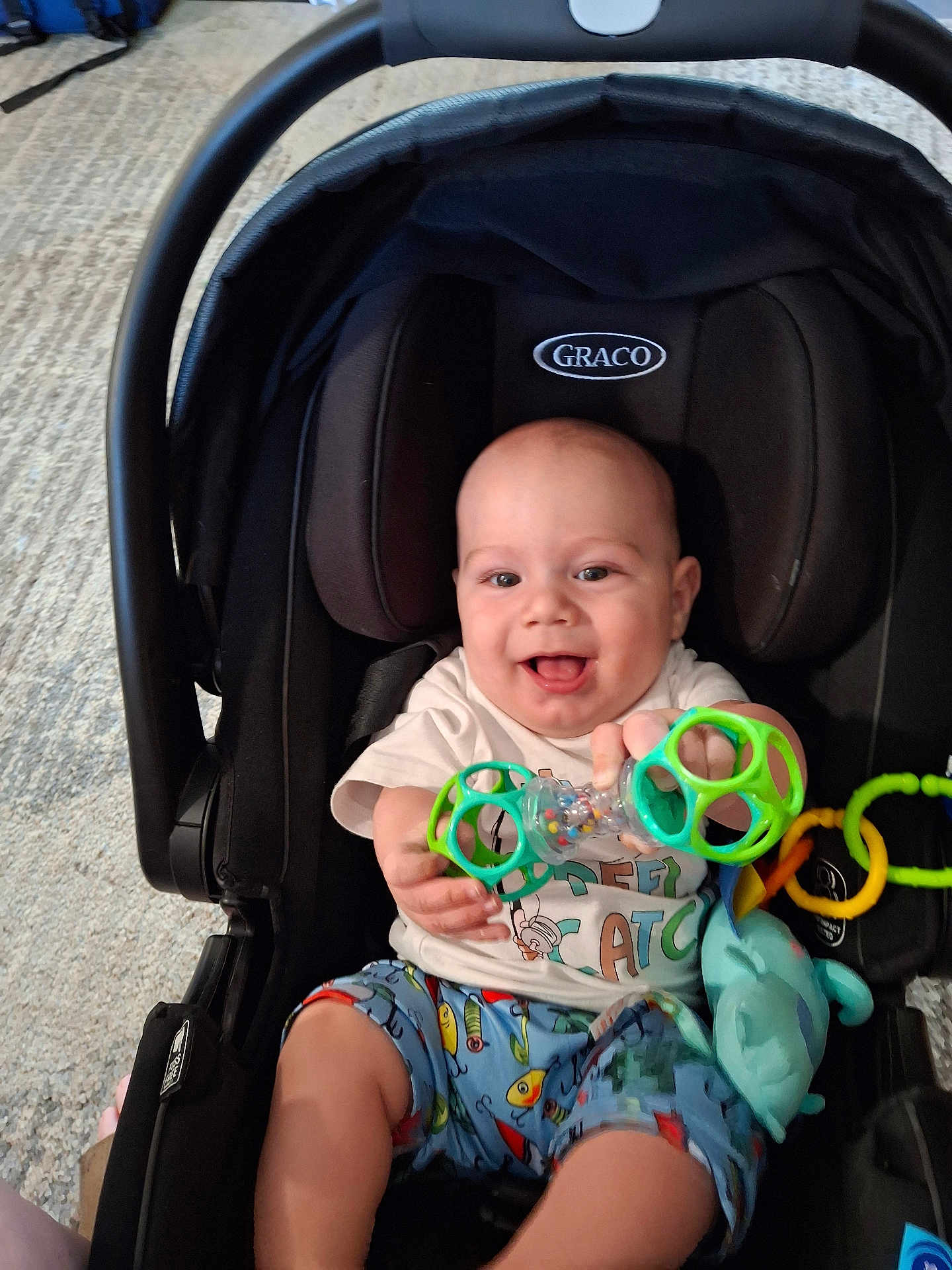 Bradley joined the competition — help win amazing prizes! baby, car_seat, toy, smiling, indoor, child, graco, clothing, shorts, white_shirt, happy, infant, seat, playful, colorful, hand, face, person, cute, young