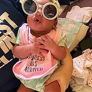 Angel is registered to the contest to win money with this photo: baby, child, sunglasses, bib, pink, person, hand, clothing, indoor, cute, infant, face, holding, accessory, fabric, blanket, pillow, skin, expression, casual