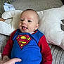 baby, blanket, blue_clothing, cozy, cute, face, hand, happy, head, indoor, infant, lying_down, person, pillow, playful, red_sleeves, smiling, soft_texture, superman_outfit, young_child