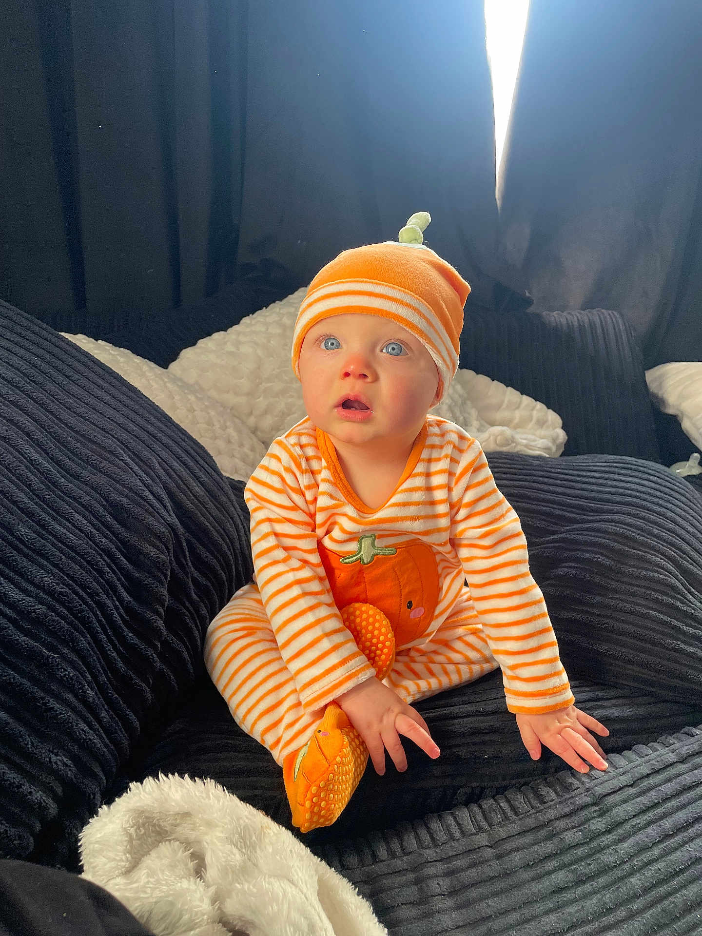 Luca is registered to the contest to win money with this photo: baby, child, infant, orange_hat, striped_clothing, pumpkin_design, blue_eyes, couch, blanket, cozy, indoor, curtains, soft_toys, sitting, cute, portrait, clothing, home, light, expression