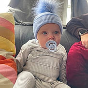 Luca joined the competition — help win amazing prizes! adult_arm, baby, blue_hat, casual, child, couch, cute, face, hat, headwear, home, indoor, infant, pacifier, person, pillow, pom_pom, sitting, striped_clothing, watch