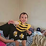 toddler, child, smiling, dress, bee_pattern, bed, blanket, pillow, indoor, person, clothing, happy, young_child, home, furniture, fabric, reaching, cute, casual, joyful