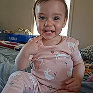 Nova is registered to the contest to win money with this photo: toddler, child, smiling, pajamas, pink_clothing, bunny_print, bed, blanket, indoor, person, furniture, window, box, cheerful, happy, cute, portrait, young_child, casual, room