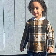 Kameron is registered to the contest to win money with this photo: child, curly_hair, plaid_shirt, jeans, standing, outdoor, sunlight, smile, fence, wooden_fence, casual_clothing, portrait, young, person, daylight, shadow, happy, front_view, clothing, nature